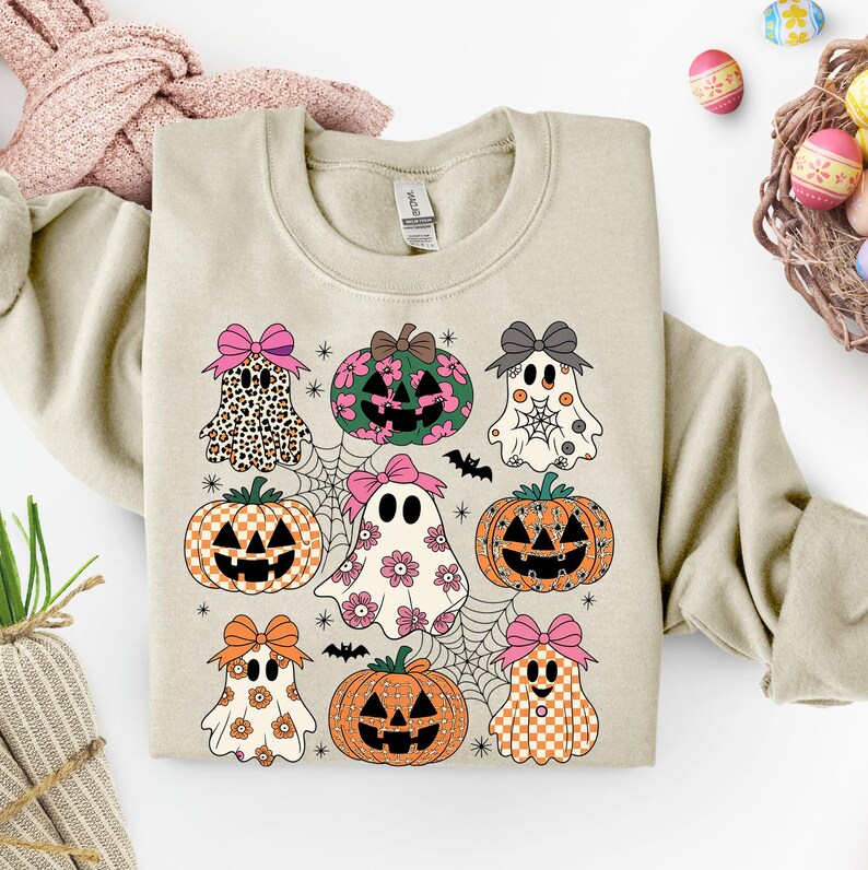 May include: Beige sweatshirt featuring a Halloween-themed design. The design includes cartoon ghosts, pumpkins, spiderwebs, and bats. The ghosts and pumpkins have bows and various patterns. The overall color palette includes orange, pink, black, and white.