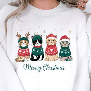 May include: White sweatshirt featuring four cartoon cats dressed in Christmas sweaters and hats. The cats are wearing red and green sweaters with paw prints and snowflake designs. The text "Meowy Christmas" is printed below the cats.