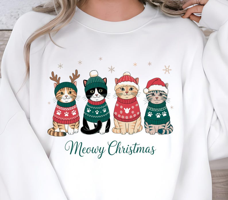 May include: White sweatshirt featuring four cartoon cats dressed in Christmas sweaters and hats. The cats are wearing red and green sweaters with paw prints and snowflake designs. The text "Meowy Christmas" is printed below the cats.