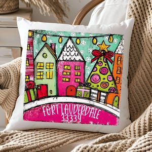 May include: White square throw pillow with a colorful illustration of houses, a Christmas tree, and gifts. The text "FORT LAUDERDALE 33334" is printed on the pillow. The design features a festive holiday theme.