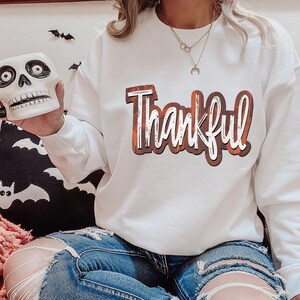 K&ouml;nnte beinhalten: Wei&szlig;er Langarm-Sweatshirt mit dem Wort "Thankful" in orangefarbenen und braunen Buchstaben bedruckt. Der Sweatshirt wird von einer Person mit einer sch&auml;delf&ouml;rmigen Tasse in der Hand getragen.