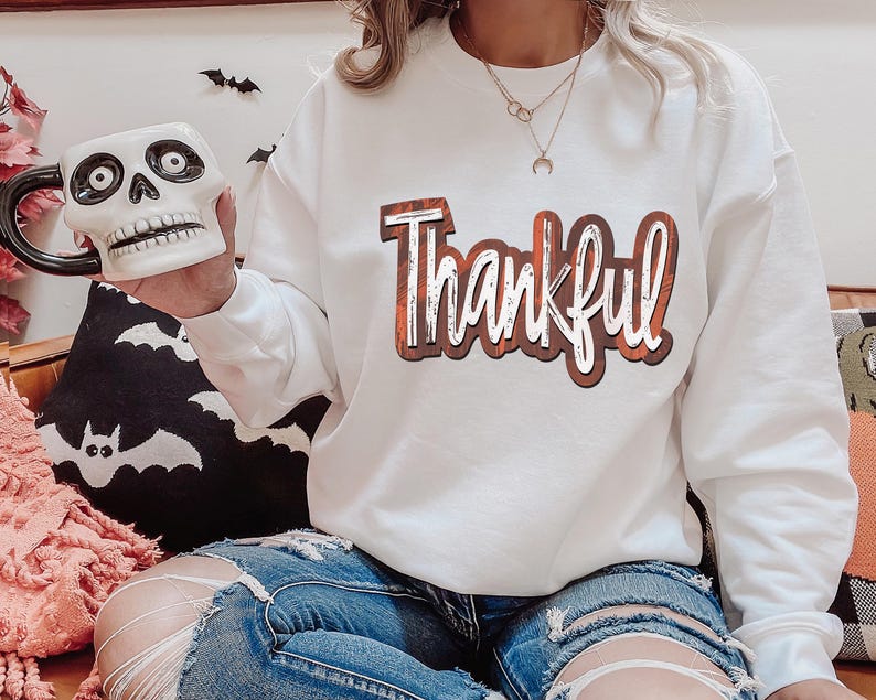 K&ouml;nnte beinhalten: Wei&szlig;er Langarm-Sweatshirt mit dem Wort "Thankful" in orangefarbenen und braunen Buchstaben bedruckt. Der Sweatshirt wird von einer Person mit einer sch&auml;delf&ouml;rmigen Tasse in der Hand getragen.