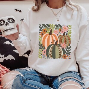 Pu&ograve; includere: Felpa bianca con una grafica di tre zucche in un design floreale. Le zucche sono verdi, arancioni e marroni su uno sfondo bianco.