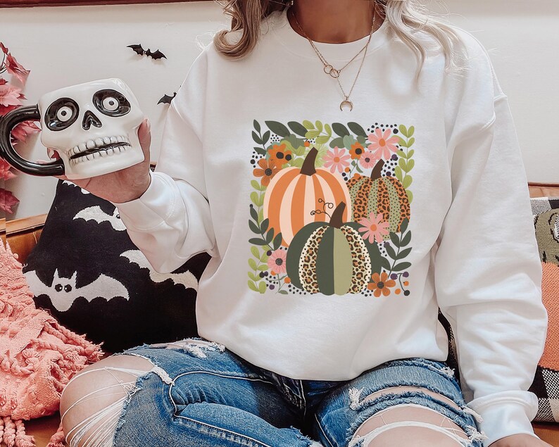 Pu&ograve; includere: Felpa bianca con una grafica di tre zucche in un design floreale. Le zucche sono verdi, arancioni e marroni su uno sfondo bianco.