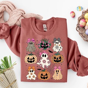 May include: A terracotta-colored sweatshirt featuring a Halloween-themed design. The design includes various pumpkins and ghosts with bows, spiderwebs, and bats. The pumpkins and ghosts have different patterns and colors, including leopard print, floral, and checkered designs.