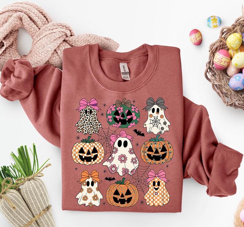 May include: A terracotta-colored sweatshirt featuring a Halloween-themed design. The design includes various pumpkins and ghosts with bows, spiderwebs, and bats. The pumpkins and ghosts have different patterns and colors, including leopard print, floral, and checkered designs.