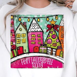 May include: White sweatshirt featuring a colorful graphic of a winter scene with houses, a Christmas tree, and gifts. The text "FORT LAUDERDALE 33334" is displayed at the bottom of the graphic. The design includes a star and a red bow.