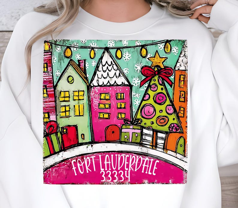 May include: White sweatshirt featuring a colorful graphic of a winter scene with houses, a Christmas tree, and gifts. The text "FORT LAUDERDALE 33334" is displayed at the bottom of the graphic. The design includes a star and a red bow.