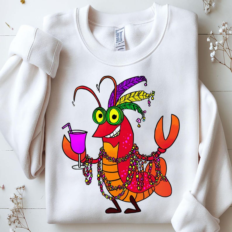 May include: White sweatshirt featuring a cartoon crawfish with a festive design. The crawfish is red, orange, and yellow, wearing a feathered headdress, green-rimmed glasses, and a beaded necklace. It holds a purple cocktail with a straw.