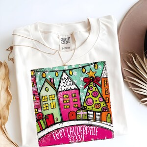 May include: Cream-colored t-shirt with a colorful graphic of a holiday scene. The design features stylized houses, a Christmas tree, and the text "Fort Lauderdale 33334". A gold necklace and a brown hat are also visible.