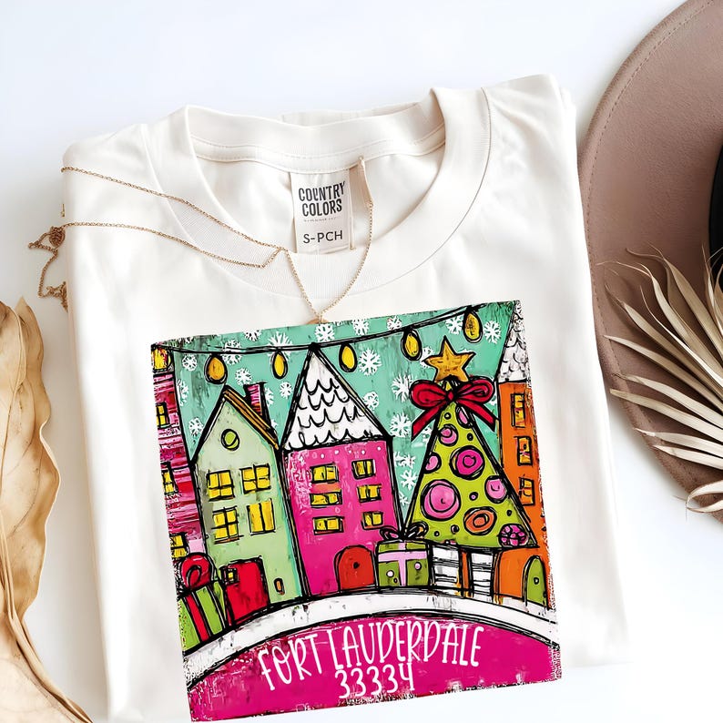 May include: Cream-colored t-shirt with a colorful graphic of a holiday scene. The design features stylized houses, a Christmas tree, and the text "Fort Lauderdale 33334". A gold necklace and a brown hat are also visible.