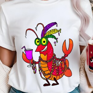 May include: White t-shirt featuring a cartoon crawfish with a festive design. The crawfish is red, orange, and yellow, wearing a feathered headdress and holding a purple drink. It is adorned with colorful beads.
