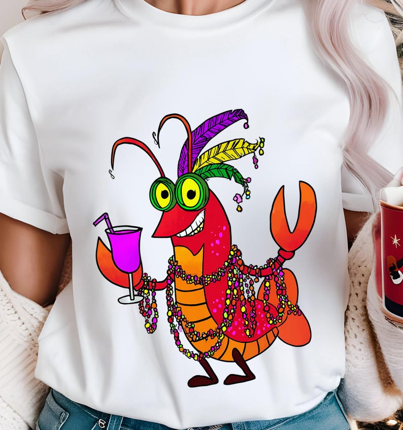 May include: White t-shirt featuring a cartoon crawfish with a festive design. The crawfish is red, orange, and yellow, wearing a feathered headdress and holding a purple drink. It is adorned with colorful beads.