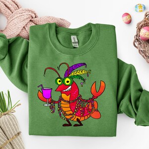 May include: Green sweatshirt featuring a cartoon crawfish with a purple drink, Mardi Gras beads, and a feathered headdress. The crawfish is red, orange, and yellow with green eyes. Easter eggs and a woven basket are in the background.