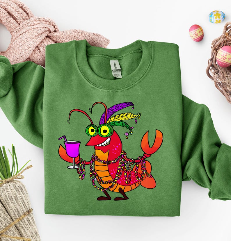 May include: Green sweatshirt featuring a cartoon crawfish with a purple drink, Mardi Gras beads, and a feathered headdress. The crawfish is red, orange, and yellow with green eyes. Easter eggs and a woven basket are in the background.