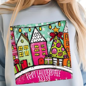 May include: Light blue sweatshirt featuring a colorful illustration of a winter scene with houses, a Christmas tree, and the text "FORT LAUDERDALE 33334". The design includes a star, snowflakes, and gift boxes.