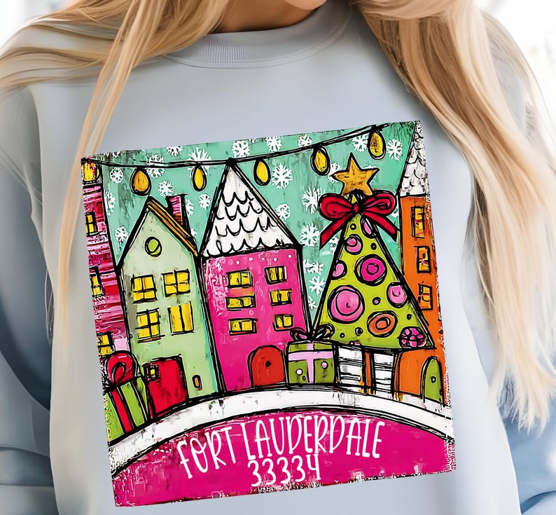 May include: Light blue sweatshirt featuring a colorful illustration of a winter scene with houses, a Christmas tree, and the text "FORT LAUDERDALE 33334". The design includes a star, snowflakes, and gift boxes.