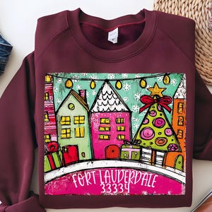 May include: Burgundy sweatshirt featuring a colorful illustration of a winter scene with houses, a Christmas tree, and the text "Fort Lauderdale 33334". The design includes gift boxes and string lights, creating a festive holiday theme.
