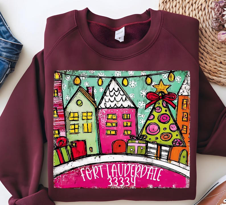 May include: Burgundy sweatshirt featuring a colorful illustration of a winter scene with houses, a Christmas tree, and the text "Fort Lauderdale 33334". The design includes gift boxes and string lights, creating a festive holiday theme.