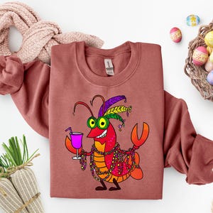 May include: A brick-red sweatshirt featuring a cartoon crawfish with Mardi Gras beads, a purple drink, and a festive headdress. The crawfish is smiling and holding a cocktail. Easter eggs are in a basket.