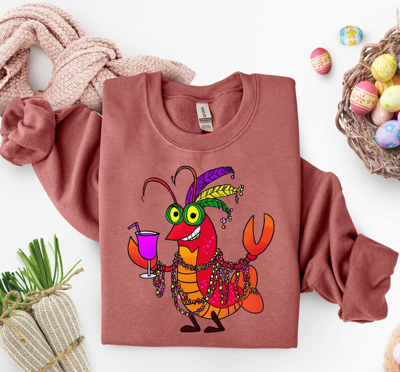 May include: A brick-red sweatshirt featuring a cartoon crawfish with Mardi Gras beads, a purple drink, and a festive headdress. The crawfish is smiling and holding a cocktail. Easter eggs are in a basket.