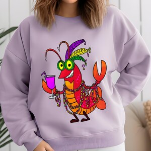 May include: A light purple sweatshirt featuring a cartoon crawfish character. The crawfish is red and orange, wearing a feathered headdress, holding a cocktail, and adorned with beaded necklaces. The design is a fun, festive graphic.