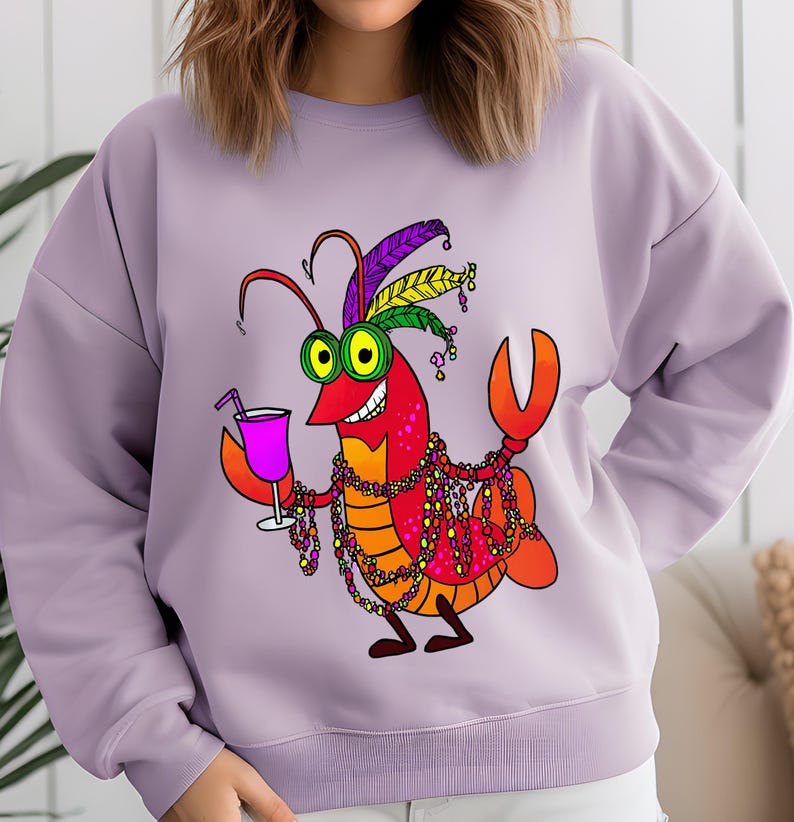 May include: A light purple sweatshirt featuring a cartoon crawfish character. The crawfish is red and orange, wearing a feathered headdress, holding a cocktail, and adorned with beaded necklaces. The design is a fun, festive graphic.