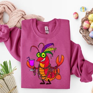 May include: A magenta sweatshirt featuring a cartoon lobster with Mardi Gras beads, a cocktail, and a feathered headdress. The lobster is in shades of red, orange, and yellow, with green eyes. Easter eggs and a basket are in the background.