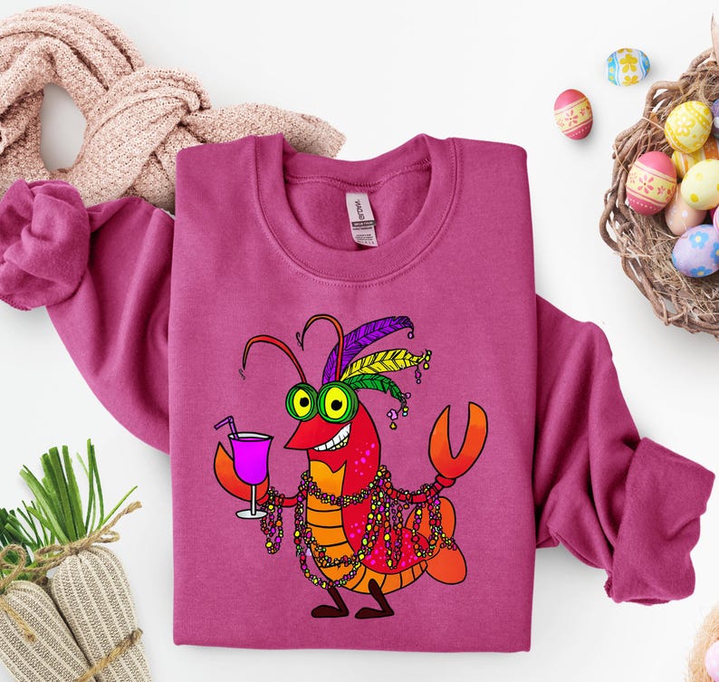 May include: A magenta sweatshirt featuring a cartoon lobster with Mardi Gras beads, a cocktail, and a feathered headdress. The lobster is in shades of red, orange, and yellow, with green eyes. Easter eggs and a basket are in the background.