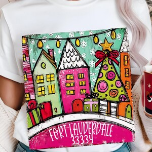 May include: White t-shirt featuring a colorful illustration of a winter scene with houses, a Christmas tree, and the text "Fort Lauderdale 33334". The design includes a pink, green, and orange color palette.