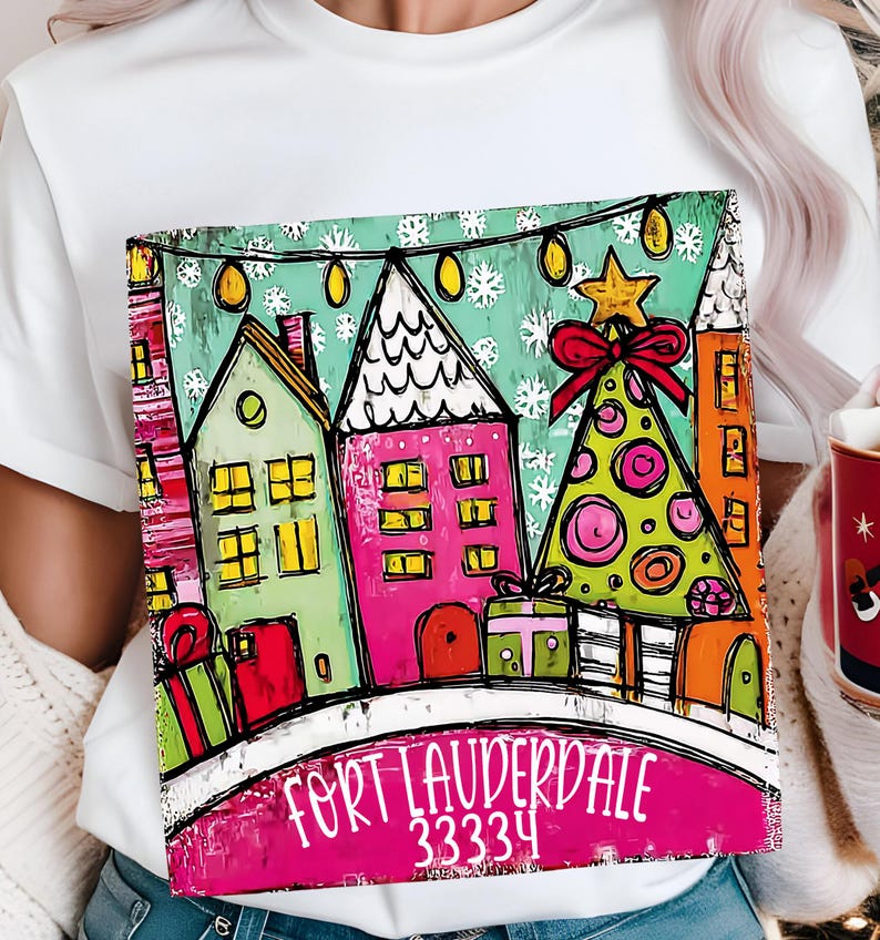 May include: White t-shirt featuring a colorful illustration of a winter scene with houses, a Christmas tree, and the text "Fort Lauderdale 33334". The design includes a pink, green, and orange color palette.