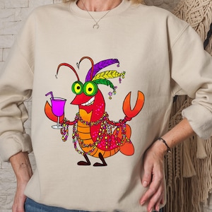 May include: Beige sweatshirt featuring a cartoon lobster with a festive Mardi Gras design. The lobster is red, orange, and yellow, wearing a feathered headdress, beads, and holding a cocktail. The design is centered on the front of the shirt.