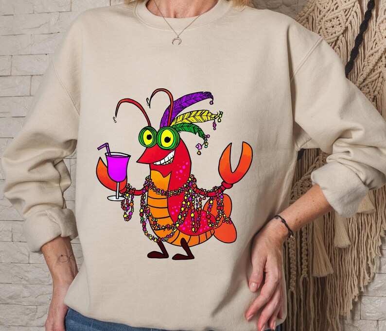 May include: Beige sweatshirt featuring a cartoon lobster with a festive Mardi Gras design. The lobster is red, orange, and yellow, wearing a feathered headdress, beads, and holding a cocktail. The design is centered on the front of the shirt.