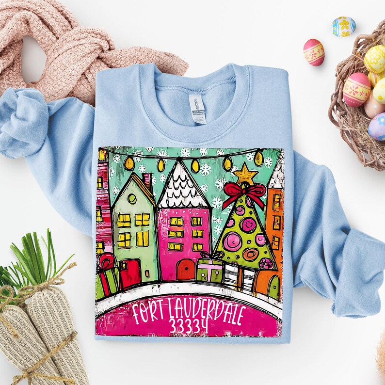 May include: Light blue sweatshirt featuring a colorful illustration of houses and a Christmas tree. The graphic includes the text "Fort Lauderdale 33334" in a pink banner. The design has a festive, whimsical style.
