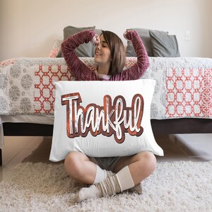 K&ouml;nnte beinhalten: Ein wei&szlig;es Kissen mit dem Wort "Thankful" in orangefarbenen und braunen Buchstaben bedruckt. Das Kissen liegt auf einem Bett, auf dem eine Frau sitzt.
