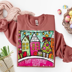 May include: A terracotta-colored sweatshirt featuring a colorful graphic of houses, a Christmas tree, and the text "FORT LAUDERDALE 33334". The design includes a red bow and star. The sweatshirt is folded with a pink scarf.