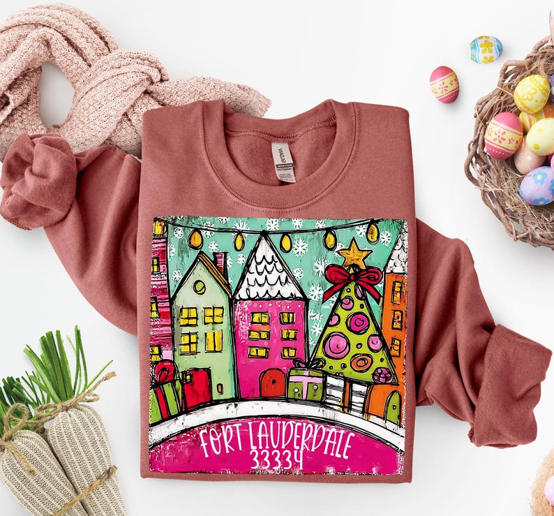 May include: A terracotta-colored sweatshirt featuring a colorful graphic of houses, a Christmas tree, and the text "FORT LAUDERDALE 33334". The design includes a red bow and star. The sweatshirt is folded with a pink scarf.