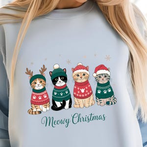 May include: Light blue sweatshirt featuring four cartoon cats in Christmas sweaters and hats. The cats are wearing red and green sweaters with paw prints and snowflake designs. The text "Meowy Christmas" is written in a script font below the cats.