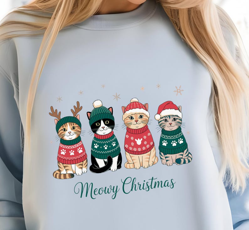 May include: Light blue sweatshirt featuring four cartoon cats in Christmas sweaters and hats. The cats are wearing red and green sweaters with paw prints and snowflake designs. The text "Meowy Christmas" is written in a script font below the cats.