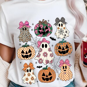 May include: White t-shirt featuring a Halloween-themed design. The design includes cartoon ghosts, pumpkins with jack-o'-lantern faces, spiderwebs, and bats. The ghosts and pumpkins have bows and floral or patterned details.