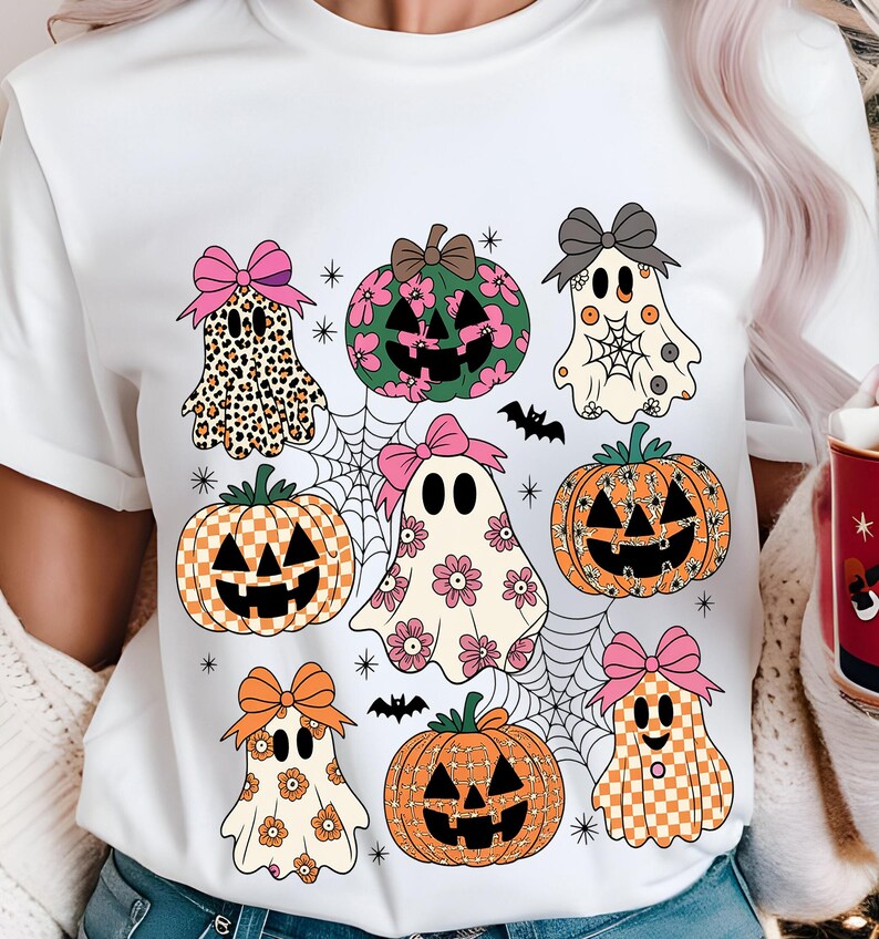 May include: White t-shirt featuring a Halloween-themed design. The design includes cartoon ghosts, pumpkins with jack-o'-lantern faces, spiderwebs, and bats. The ghosts and pumpkins have bows and floral or patterned details.