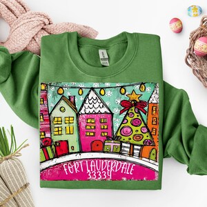 May include: Green sweatshirt featuring a colorful graphic of houses, a Christmas tree, and the text "FORT LAUDERDALE 33334". The graphic includes a string of lights and a star on top of the tree. The design is on a white background.