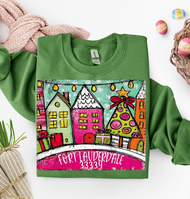 May include: Green sweatshirt featuring a colorful graphic of houses, a Christmas tree, and the text "FORT LAUDERDALE 33334". The graphic includes a string of lights and a star on top of the tree. The design is on a white background.