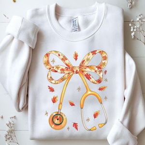 May include: White sweatshirt featuring a fall-themed design. A bow with floral patterns and a stethoscope in shades of orange and yellow are the focal point. Scattered around are red and orange leaves and star accents, creating a seasonal aesthetic.