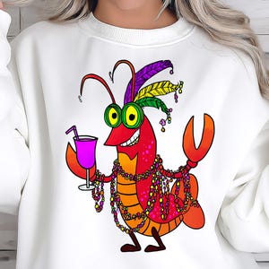 May include: White sweatshirt featuring a cartoon crawfish with a cocktail, beads, and a feathered headdress. The crawfish is red, orange, and yellow with green eyes. The beads are multi-colored, and the drink is purple.