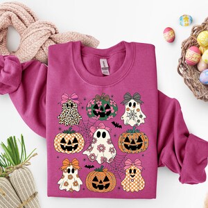 May include: A magenta sweatshirt featuring a Halloween-themed design. The design includes cartoon ghosts, pumpkins, and bows. The ghosts have floral and checkered patterns. The pumpkins have jack-o'-lantern faces. The design is set against a backdrop of spiderwebs and bats.