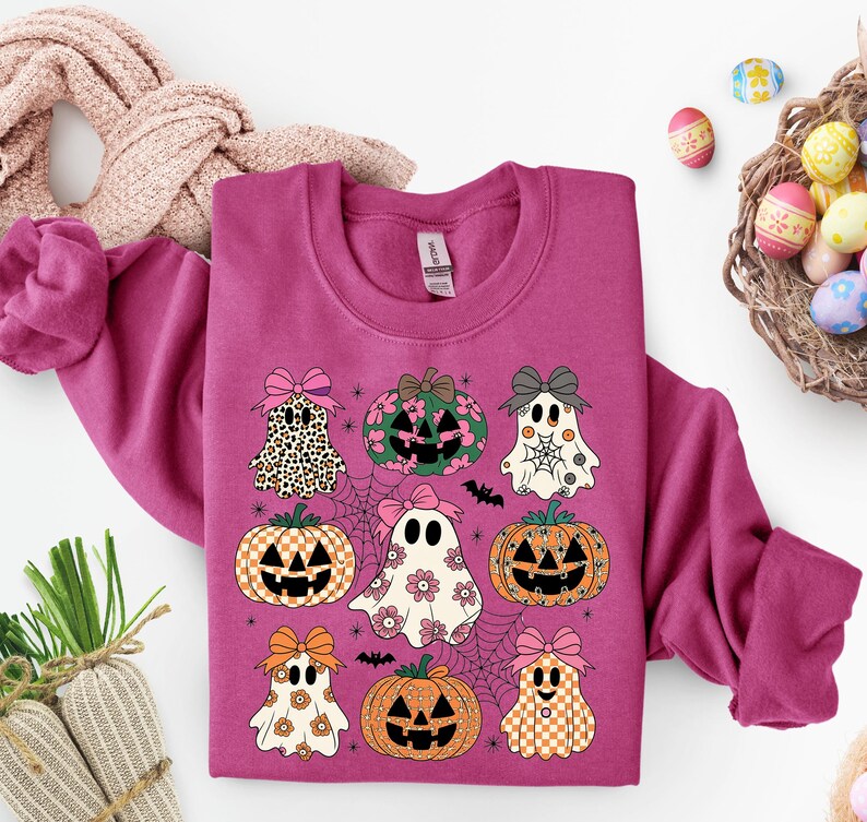 May include: A magenta sweatshirt featuring a Halloween-themed design. The design includes cartoon ghosts, pumpkins, and bows. The ghosts have floral and checkered patterns. The pumpkins have jack-o'-lantern faces. The design is set against a backdrop of spiderwebs and bats.