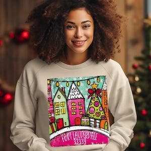 May include: Beige sweatshirt featuring a colorful Christmas-themed graphic. The graphic depicts a row of houses and a Christmas tree, with the text "FORT LAUDERDALE 33334" at the bottom. The design includes vibrant colors and festive elements.