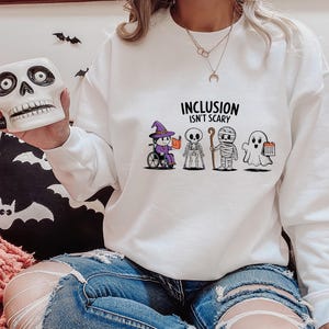 May include: White sweatshirt with the text "Inclusion Isn't Scary" and a graphic of five Halloween characters. The characters include a witch, a skeleton, a mummy, a ghost, and a ghost holding a calendar.