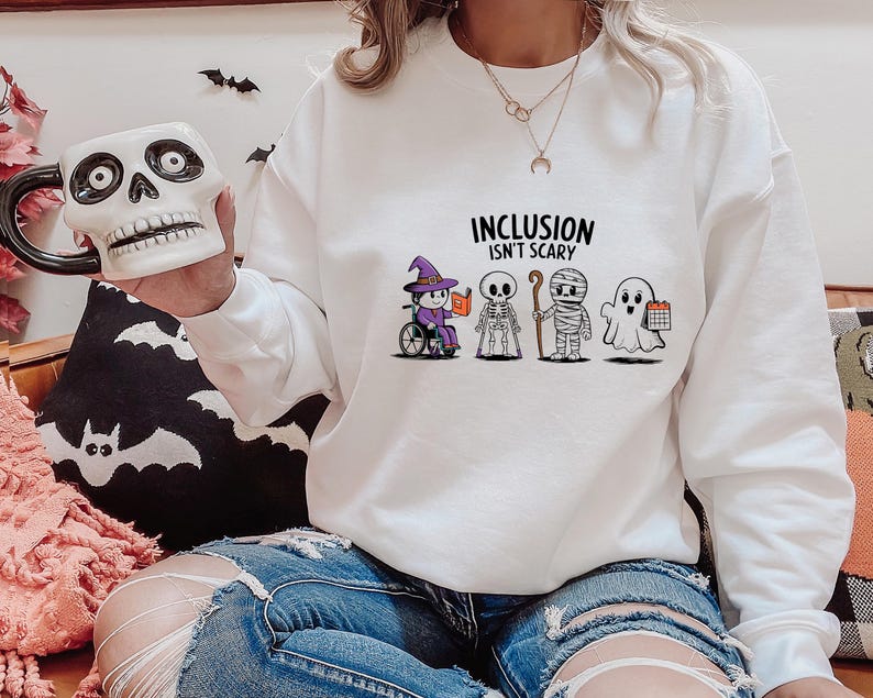 May include: White sweatshirt with the text "Inclusion Isn't Scary" and a graphic of five Halloween characters. The characters include a witch, a skeleton, a mummy, a ghost, and a ghost holding a calendar.